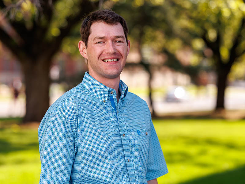Daniel Adamson headshot with a backdrop of green grass and trees. He has brown hair and is wearing a light blue short-sleeved, collared shirt.