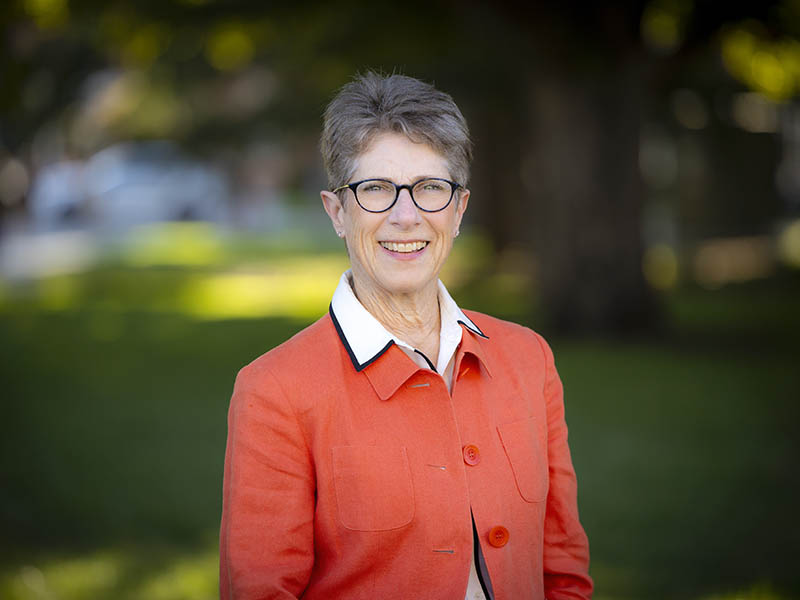 Deana Hildebrand with glasses and pixie-cut light brown hair wearing an orange dress jacket and standing in front of a blurred grove of trees.