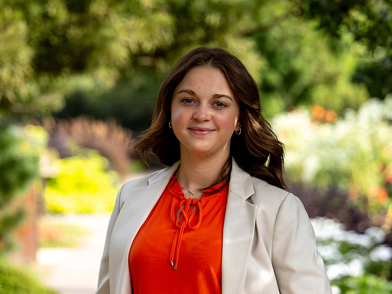 Emma Lewis standing in front of a grove of trees wearing a tan blazer and an orange shirt. She has brown shoulder-length hair.