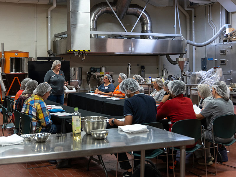 A group of men and women sit around long tables in the spacious food preparation area of the Robert M. Kerr Food and Agricultural Products Center. They are wearing hair nets and watching their breadmaking instructor at the front of the room as she stands to present instructions for a course on breadmaking.