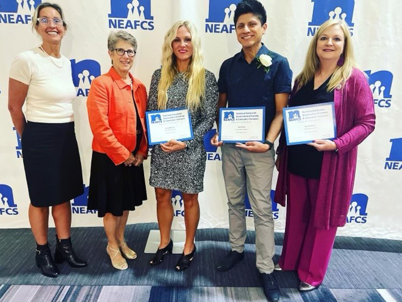 A group of five OSU Family and Consumer Sciences specialists stand in front of a conference backdrop holding paper awards in their hands