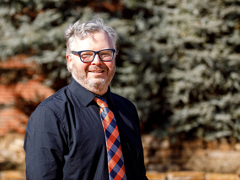 Jason Harmon stands in front of a green and brown background against an orange brick wall. He's wearing black glasses, a black, collared shirt and a blue and orange tie.