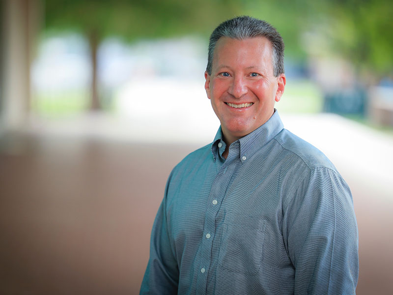 Rodney Holcomb headshot. He is wearing a light blue, long-sleeved, collared, button-up shirt. The background is blurred.