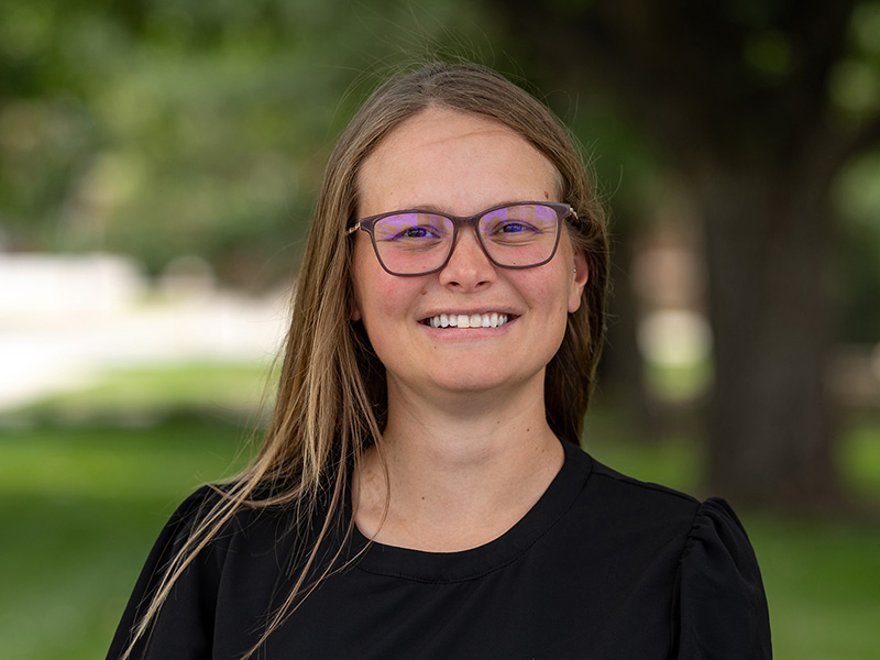 Jenny Dudak in a black shirt and glasses. Her light brown hair is down, and the background is green, blurred trees.