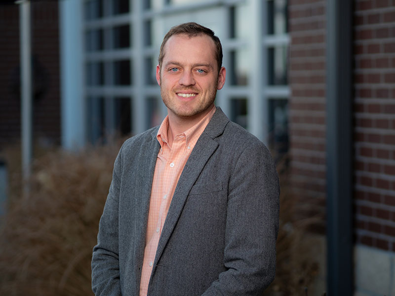 headshot of Josh Campbell with brown hair, wearing an orange collared shirt and a gray blazer