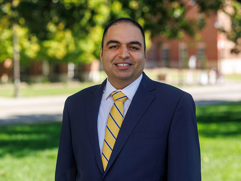 KJ Jafarzadegan stands in front of a blurred line of trees. He's wearing a navy blue suit with a yellow and blue striped tie and a light blue button up collared shirt. KJ has dark hair and eyebrows.