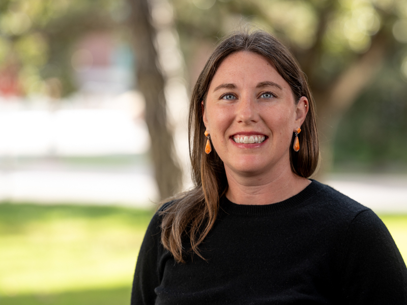 Liberty Galvin headshot, wearing a black shirt and orange earrings with her brown hair down past her shoulders.