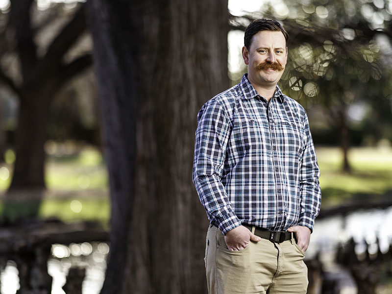 Mark Turner stands in front of a blurred large tree wearing khaki pants, a brown belt and a blue and green plaid, long-sleeved, collared shirt. He has brown hair and a dark red mustache.