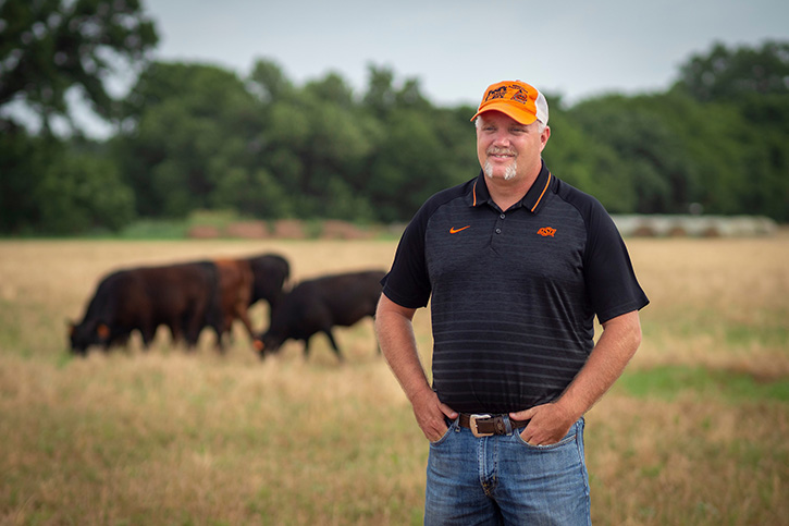 Justin Talley is standing in a field of dried, brown grass with cattle behind him in the distance. He is wearing a black OSU polo shirt, jeans and an orange OSU hat.