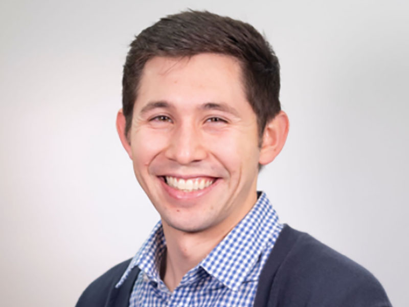 Headshot of Ty Gregson with brown hair, wearing a blue and white checkered collared shirt and blue sweater