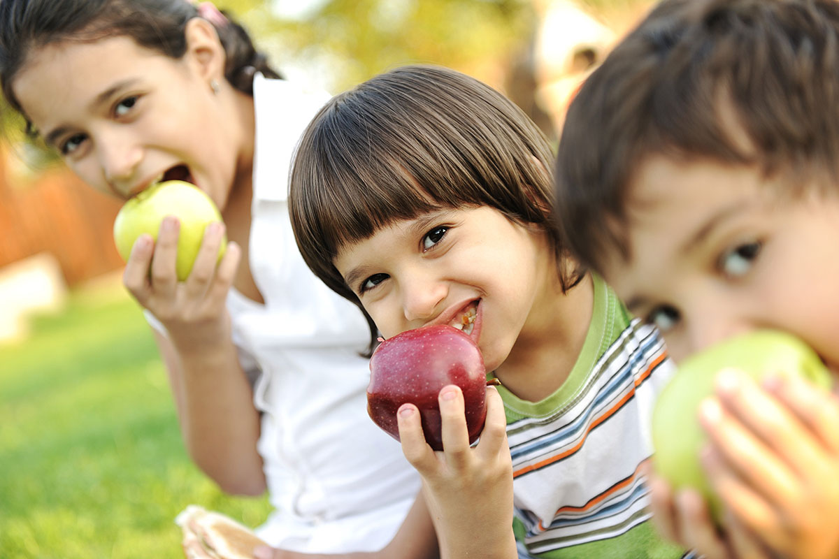 Three children are sitting down eating apples and smiling.