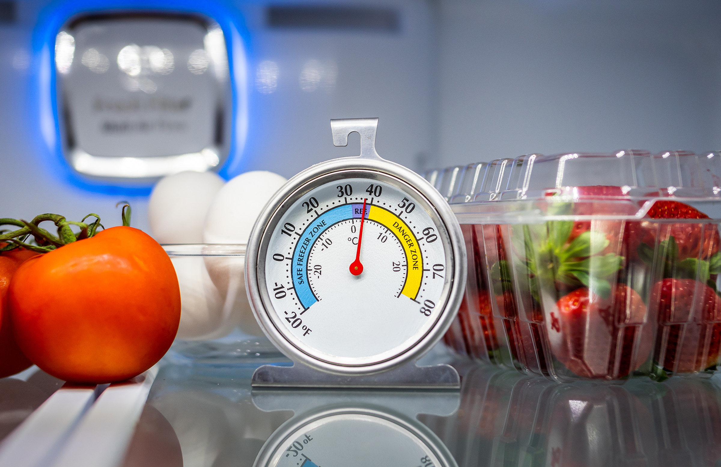 A thermometer sits on the top shelf in a refrigerator next to tomatoes, eggs and strawberries.