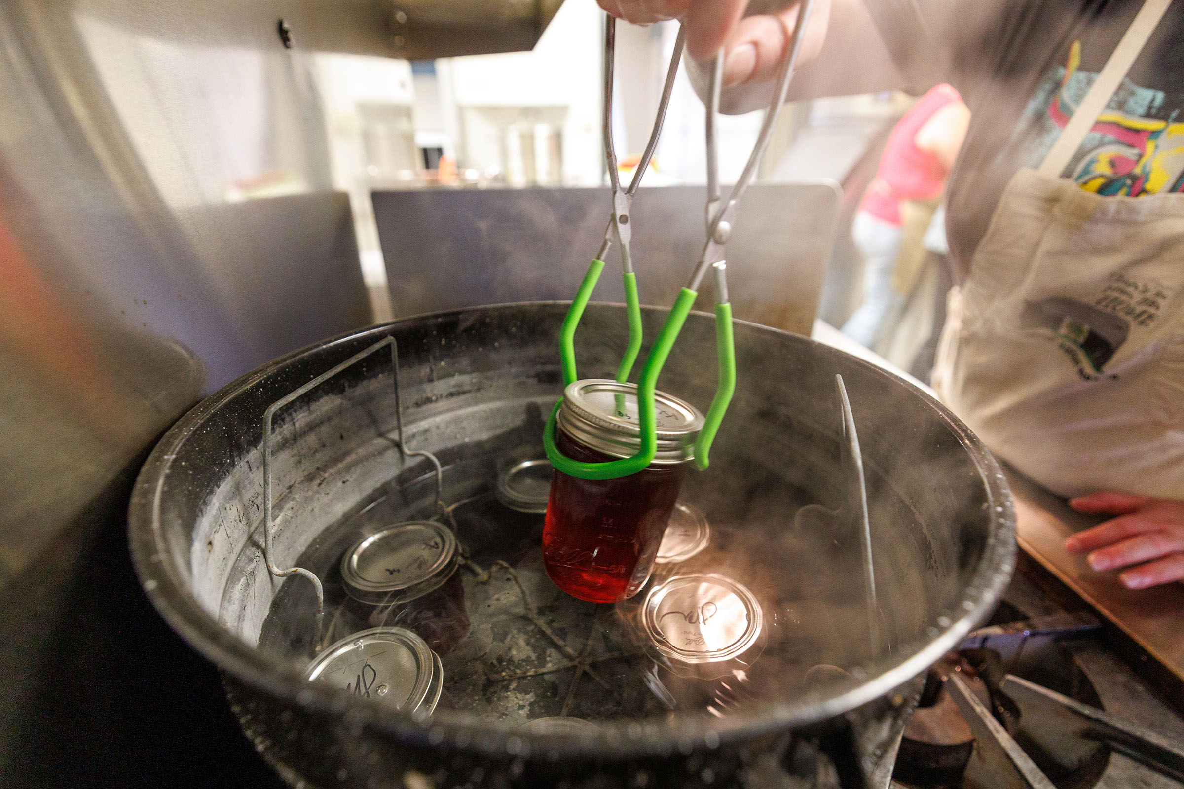 A person using green tongs to set a canned jar filled with liquid inside of a boiling pot with other canned jars.