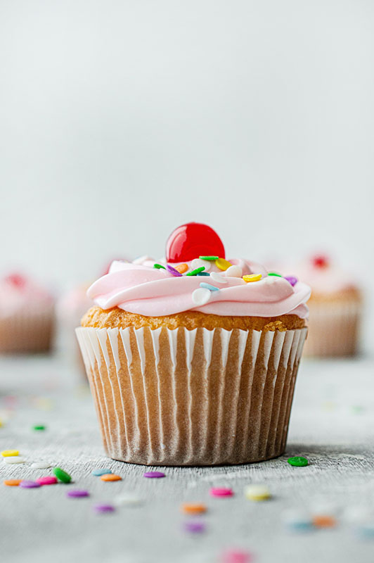 A cupcake with pink frosting and sprinkles with a red cherry on top.