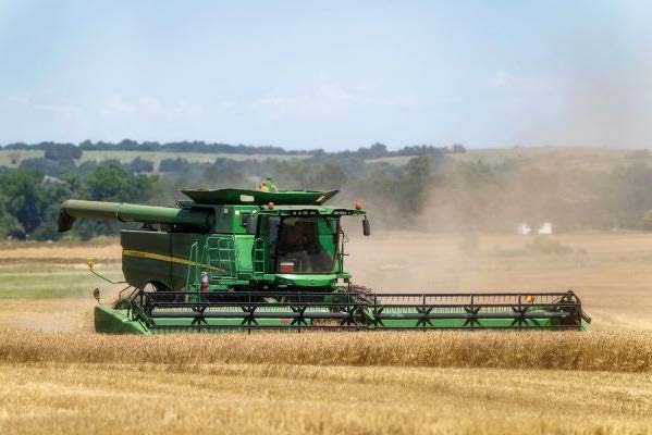 Green combine harvester harvesting a golden grain field, creating dust as it cuts crops across a broad rural landscape.