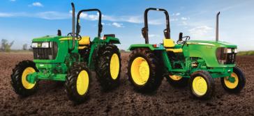Two green tractors with yellow wheels parked side by side in a freshly tilled field under a clear blue sky.