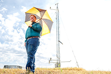 Aaron Long stands in a dry grassy field holding a large orange-and-black umbrella, with a weather monitoring station behind him under a bright, partly cloudy sky.
