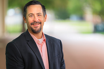 Dr. Brian Arnall wears a dark blazer and a coral button-down shirt, standing outdoors, with a softly blurred green landscape and walkway in the background.