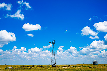 A wide-open grassland under a vivid blue sky filled with scattered white clouds, featuring a tall windmill and a small water tank on the flat horizon.