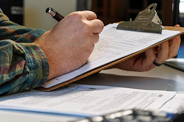 A person in a plaid shirt fills out tax forms on a clipboard at a desk, with additional paperwork and a calculator nearby.