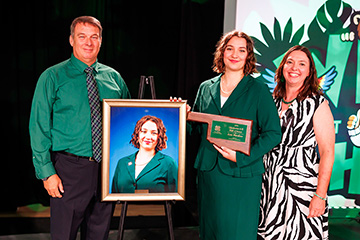2026 Oklahoma 4-H Hall of Fame inductee Kate Wardlaw stands with a plaque and her framed portrait between her parents.