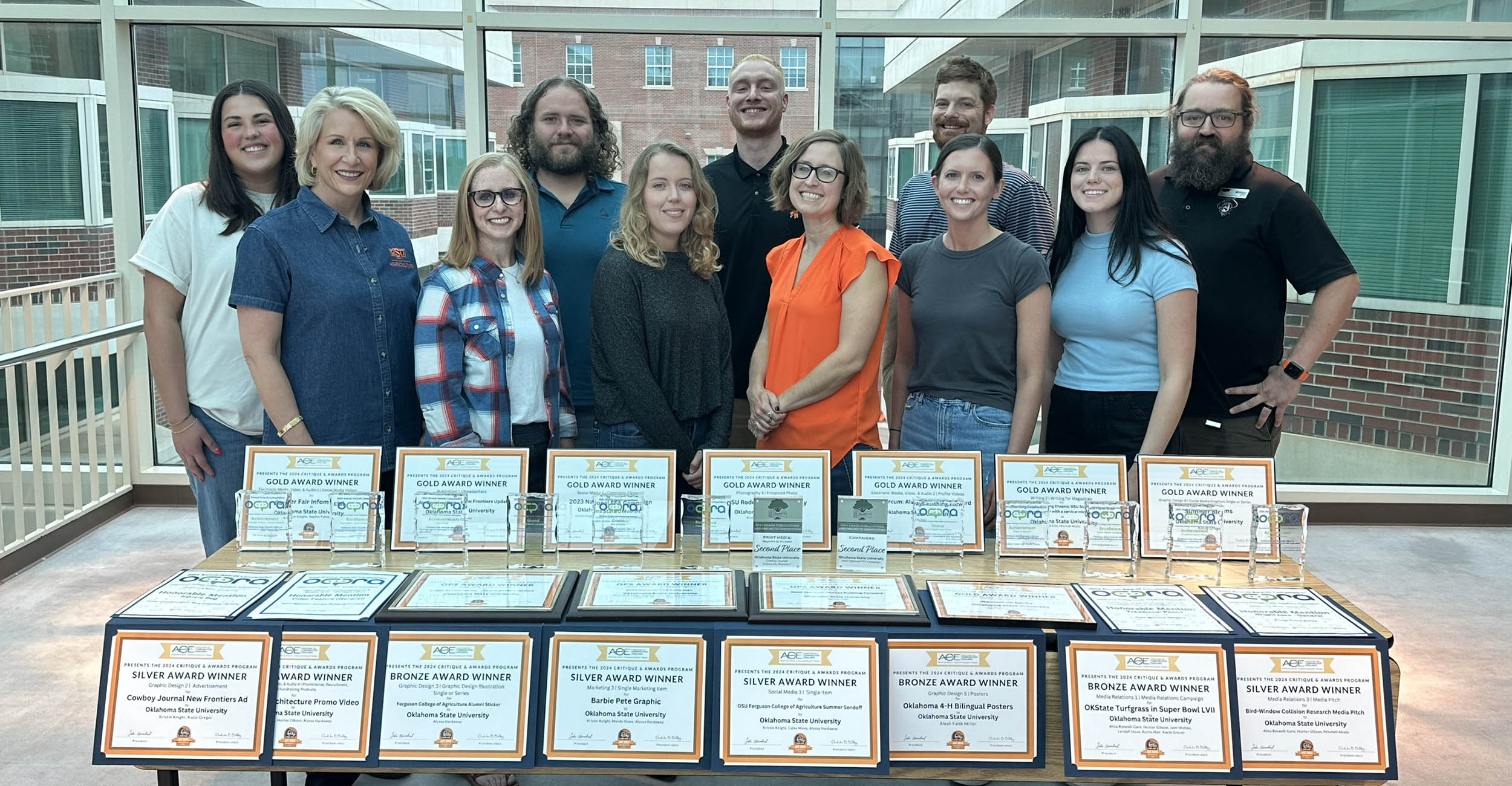 A group of 11 men and women stand in front of a table displaying plaques and certificates. The room in the background is made of glass walls.