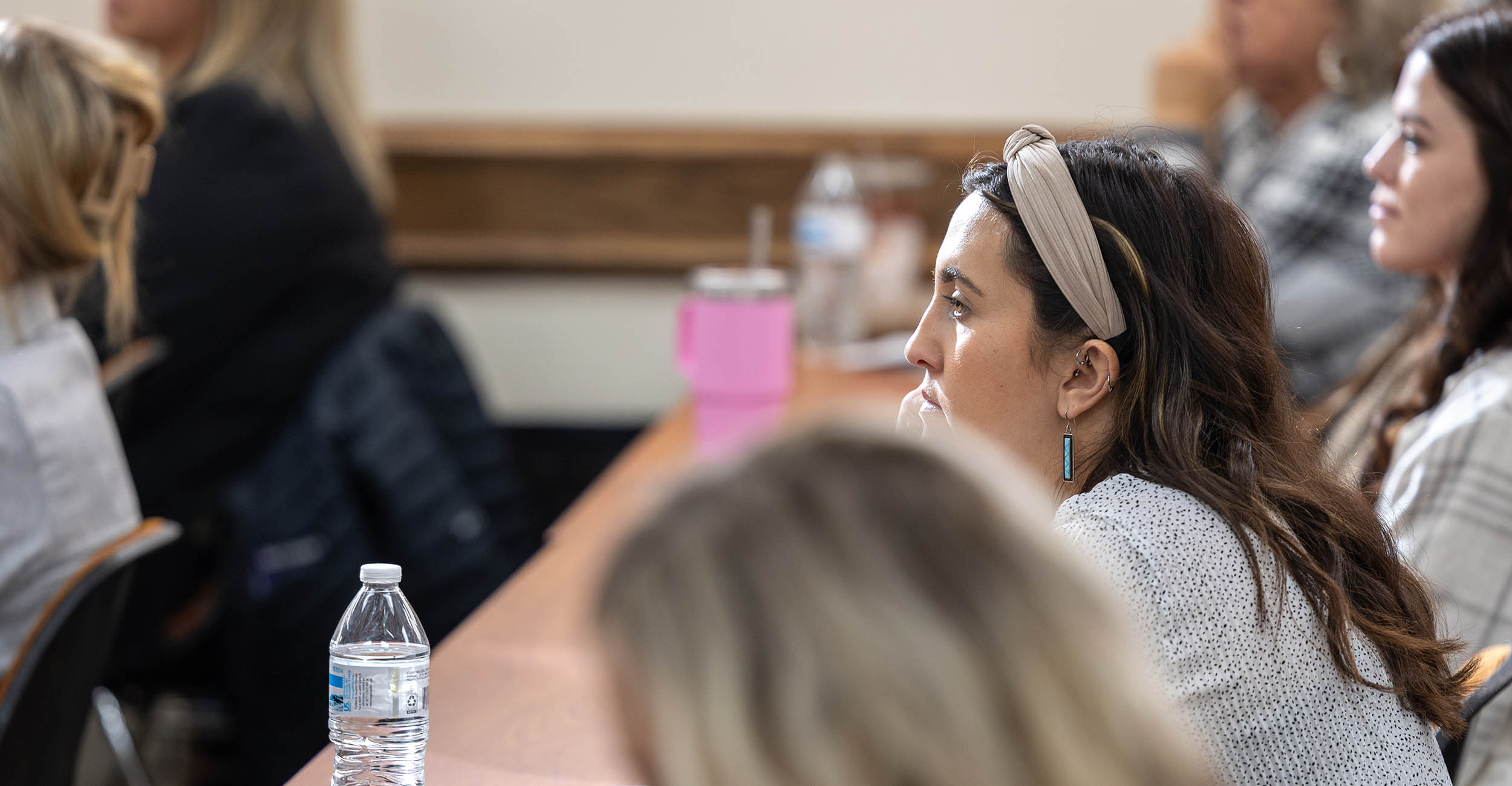 A young woman sits at a long table in a conference room, looking ahead, listening to a presentation at the front of the room. The image is of her side profile. She is wearing a tan headband and has long, brunette hair. Other women sit beside and in front of her.
