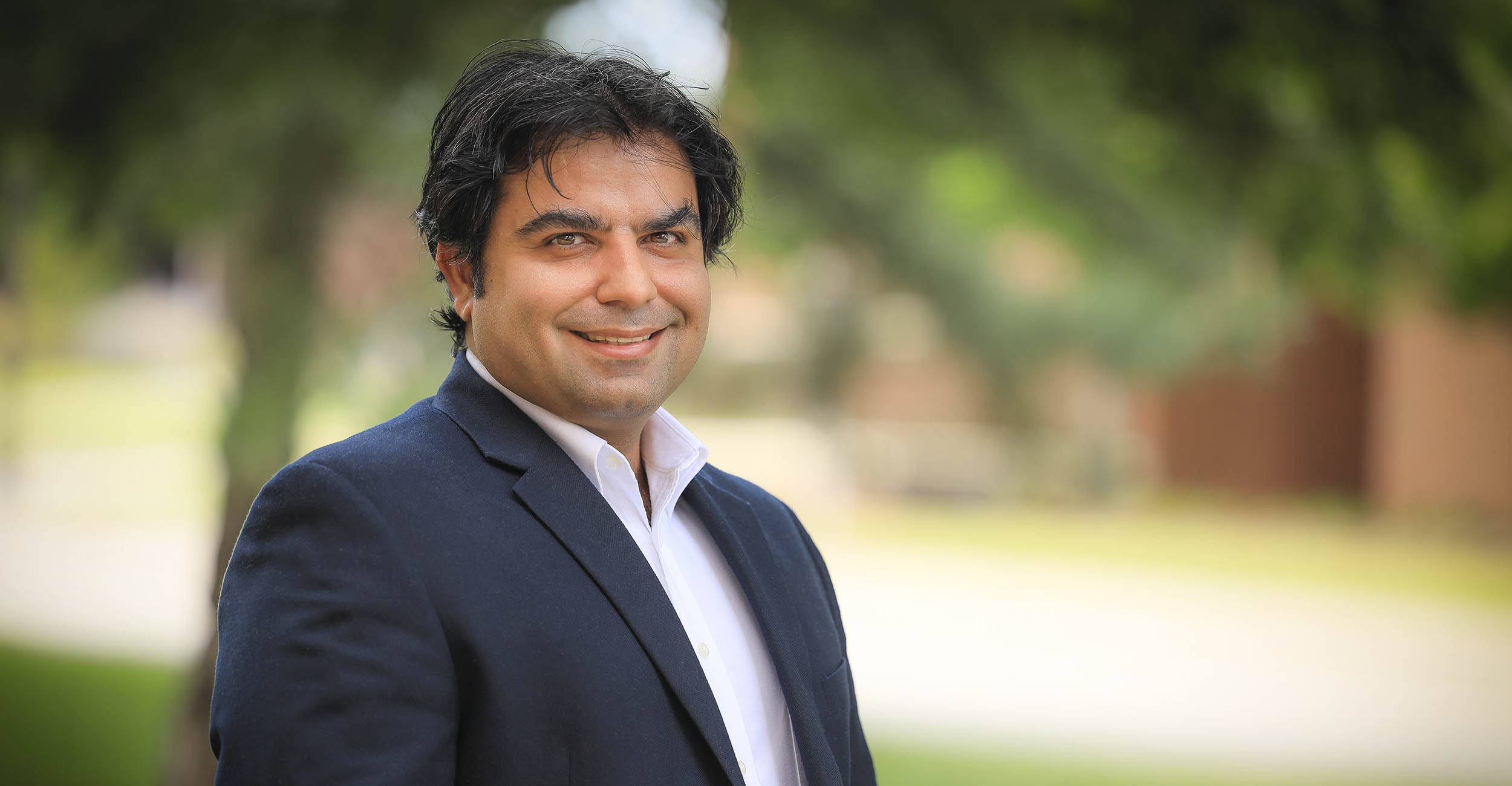 Headshot of Ali Mirchi wearing a blue suit coat and light purple collared shirt.