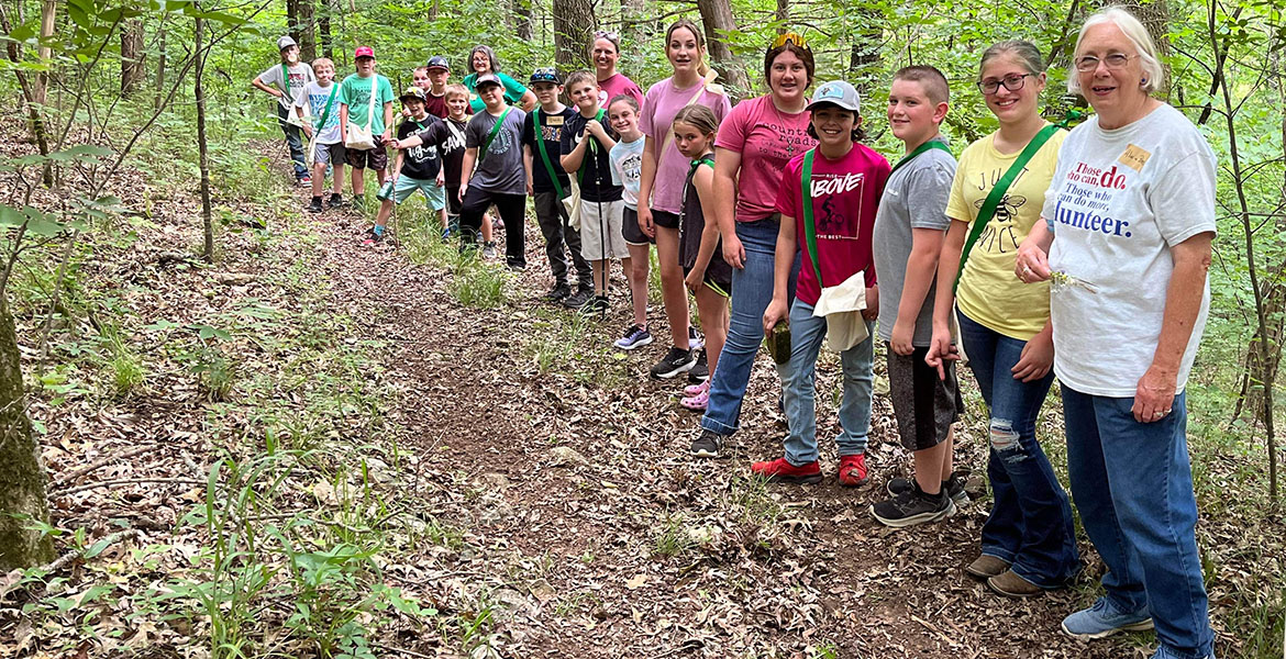 A group of people of all ages in colorful summer clothing hike in a wooded area.