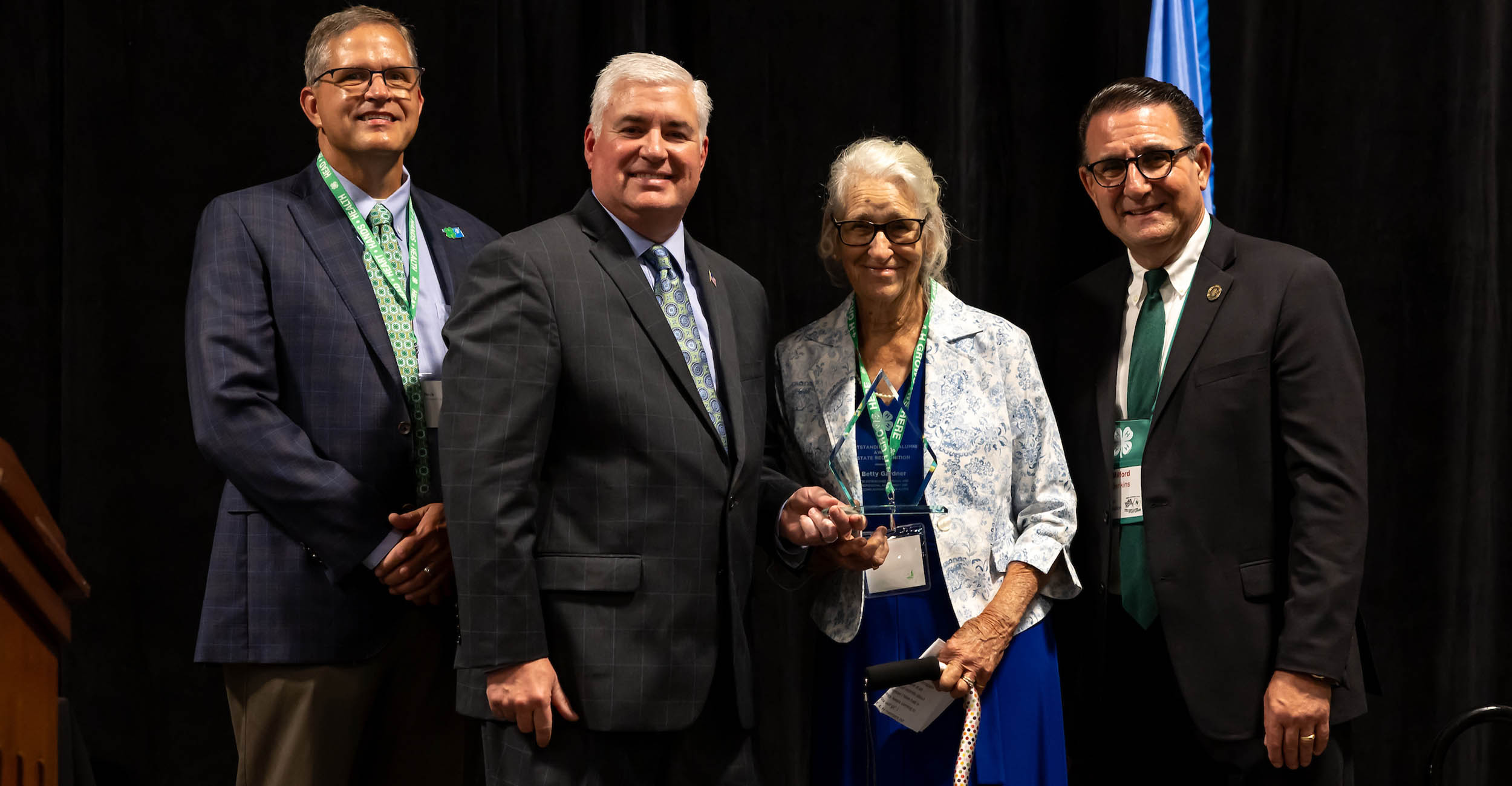 Betty Gardner stands with a group of three men in suits. She is holding a glass, clear plaque for her Oklahoma 4-H Outstanding Alumni Award. Betty has white hair and is wearing a blue dress and gray and white blazer.