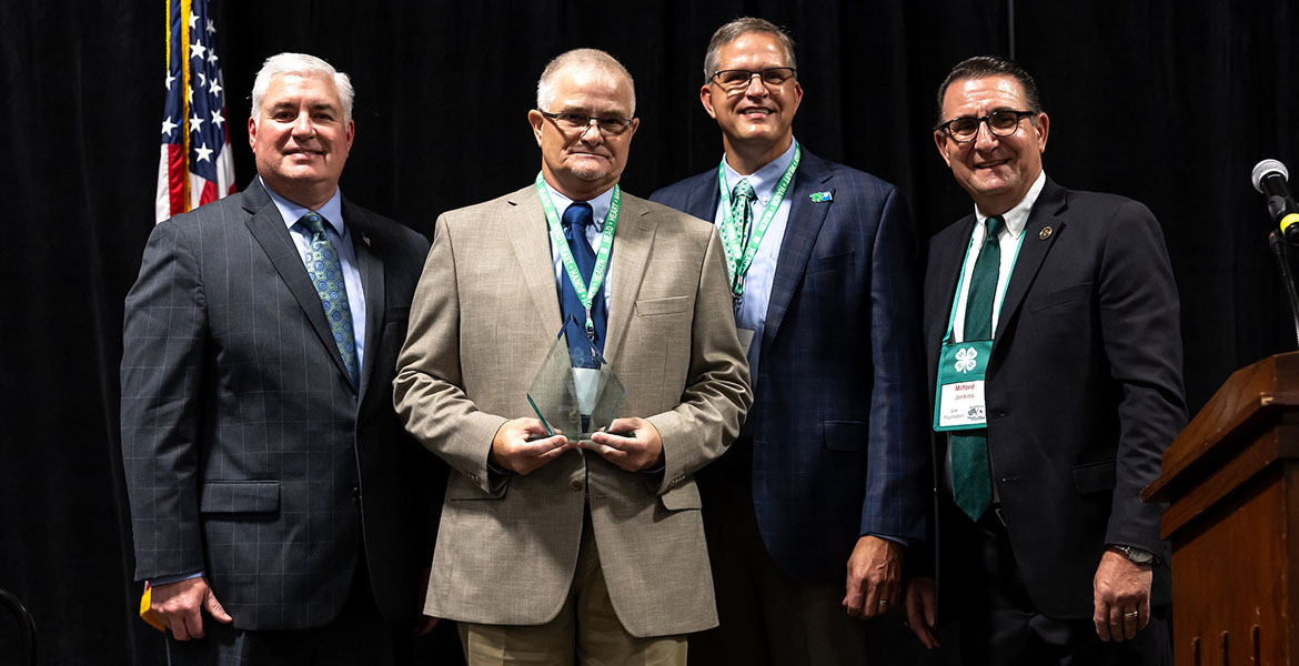 Billy stands holding his clear, glass plaque award while posing in a picture with three other men in suits. Billy is wearing glasses, a tan suit and a navy tie.