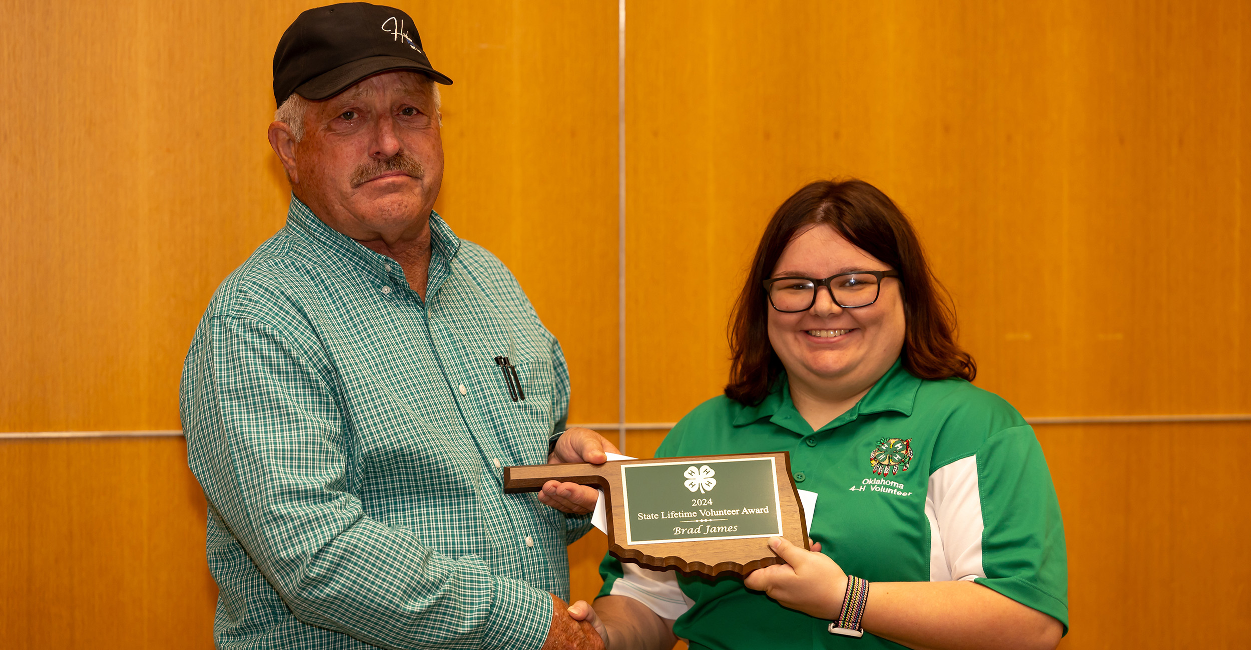 Man in green shirt and baseball cap receives an Oklahoma-shaped plaque from a woman in a green shirt who is wearing glasses.