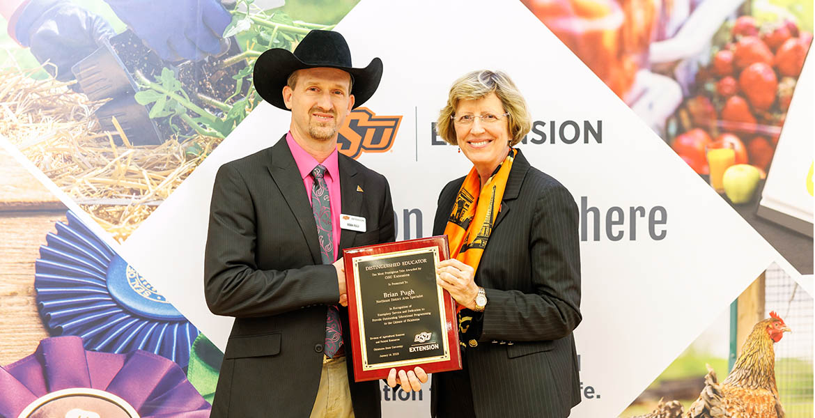 Brian Pugh receives a plaque from Damona Doye. Brian is wearing a pink shirt, pink and black tie, black suit coat and black cowboy hat. Damona is wearing a black blazer and orange scarf.