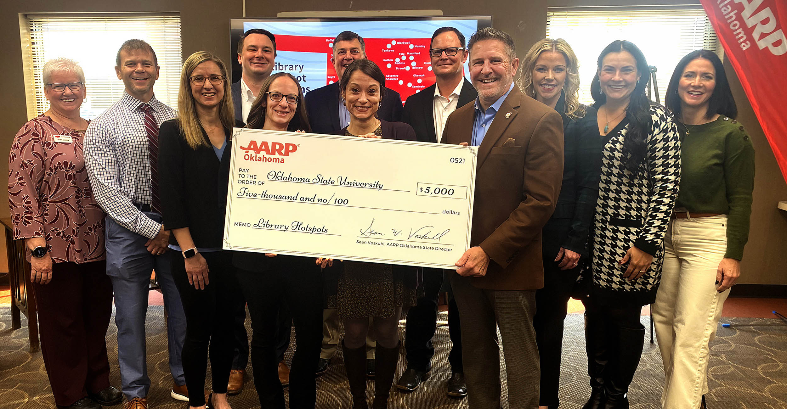 A group of men and women stand in two rows in front of a large television screen. The front three women and man hold a large presentation check addressed to Oklahoma State University in the amount of $5,000. The memo line says Library Hotspots.