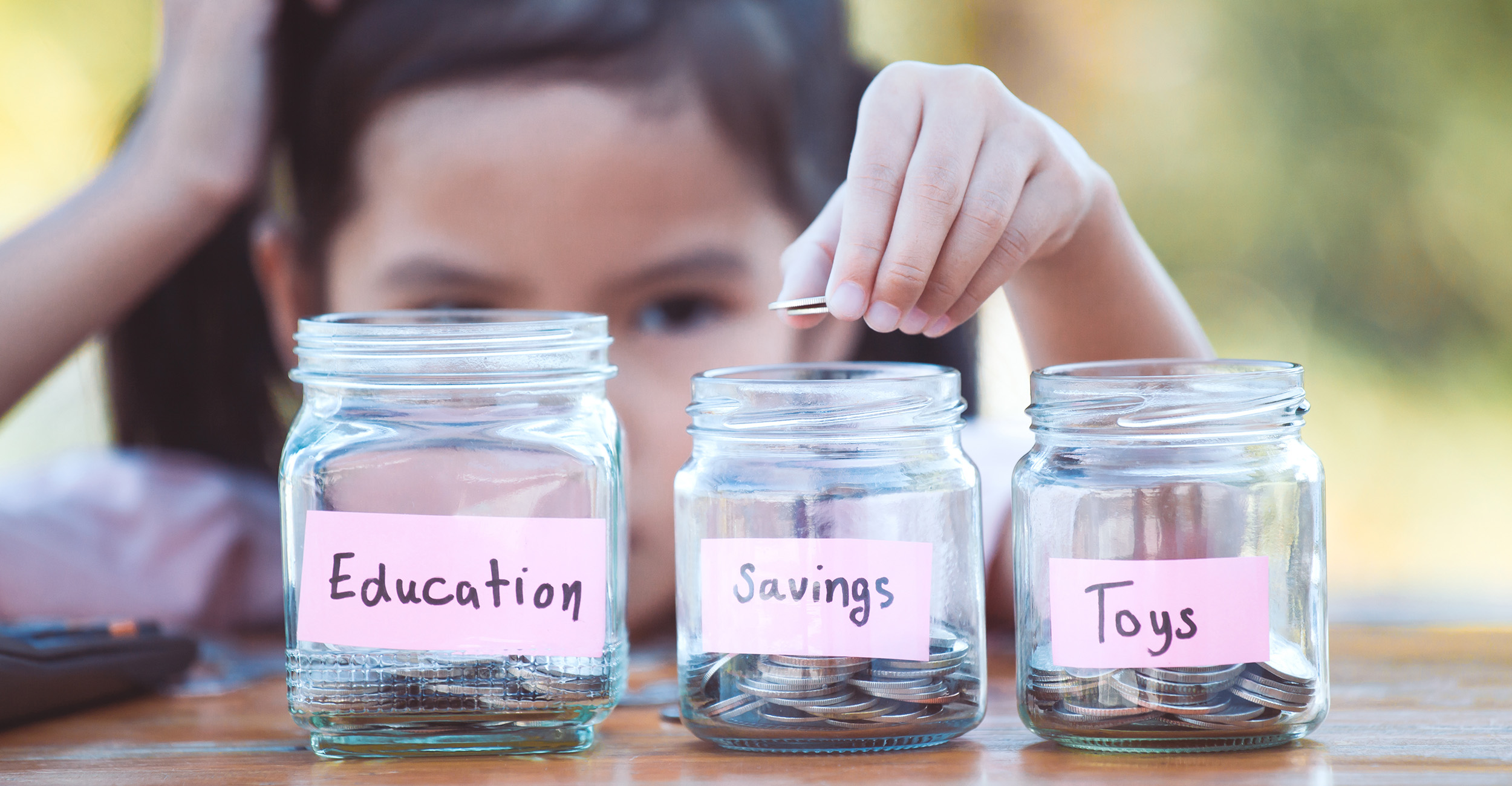 Young Asian girl dropping coins into three different clear glass jars labeled education, savings and toys.
