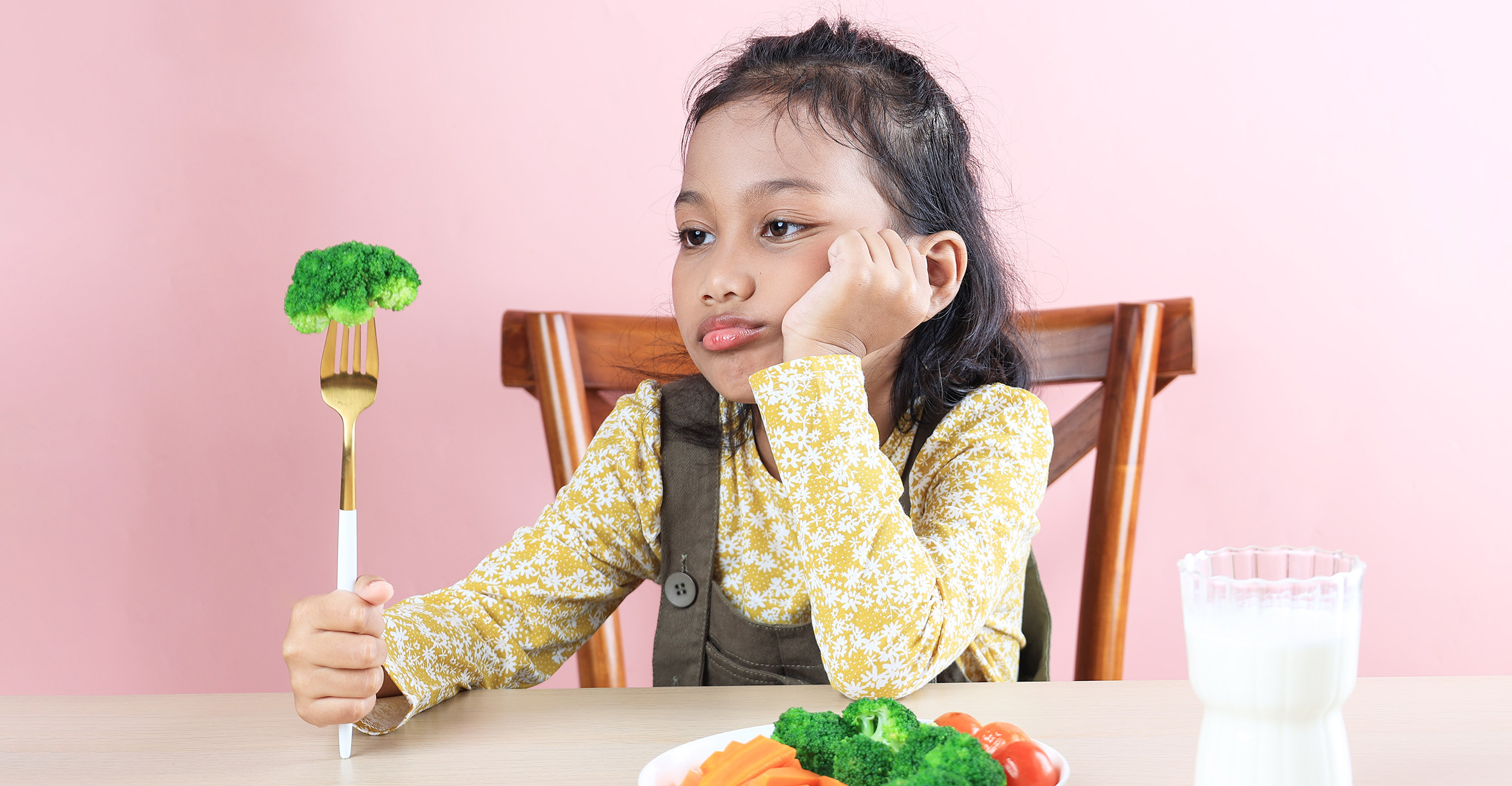 Young girl with dark hair, wearing a long-sleeved yellow shirt with white flowers under a pair of brown overalls, looks at a piece of broccoli on a fork.