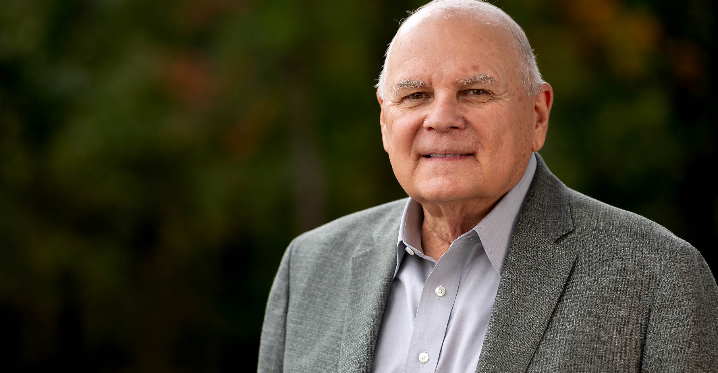 Headshot of Claude Bess wearing a gray sport coat and light blue collared, button-up shirt. The background is blurred dark green trees.
