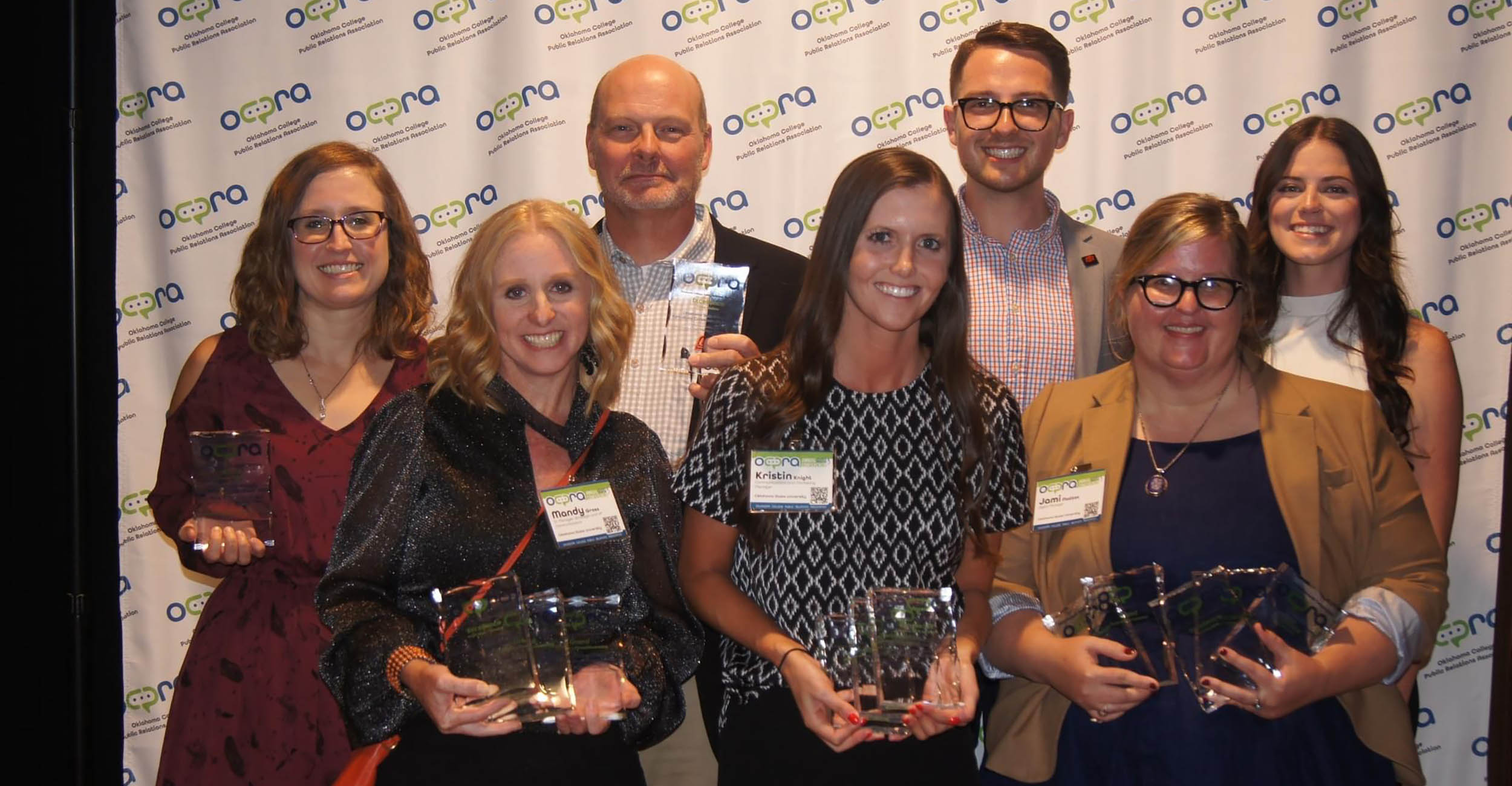 group of staff members standing in two rows holding clear, glass awards in front of a branded backdrop for the Oklahoma College Public Relations Association