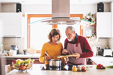 Two older adults prepare a meal together in a bright, modern kitchen. One person stirs a pot on the stovetop while the other adds chopped vegetables from a cutting board, with fresh produce and seasonings on the counter, illustrating cooking for one