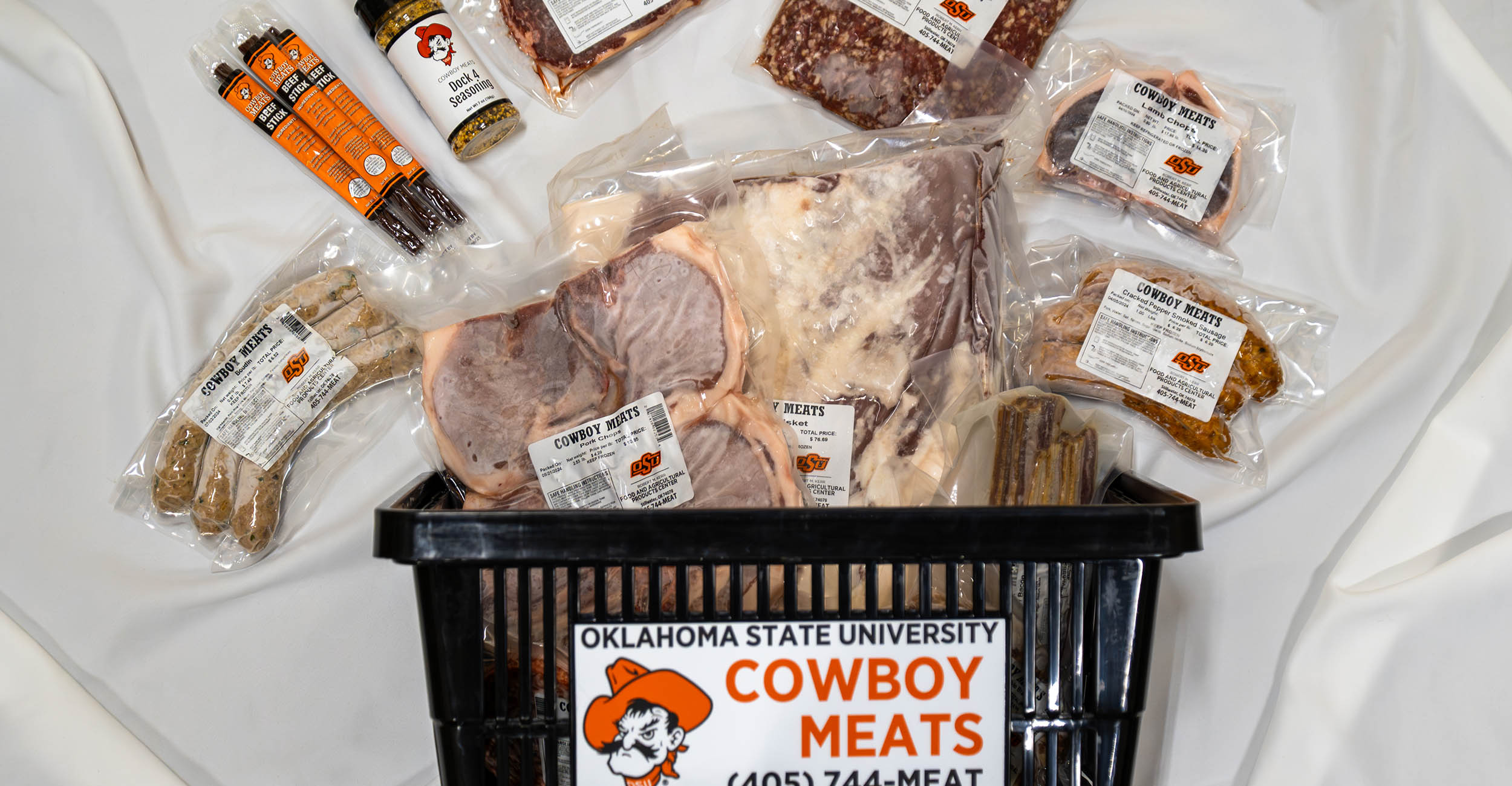 A small black grocery handheld basket with the Cowboy Meats logo on it lays on a surface. Displayed around the basket are different packaged beef, pork and lamb products prepared in the Food and Agricultural Products Center.