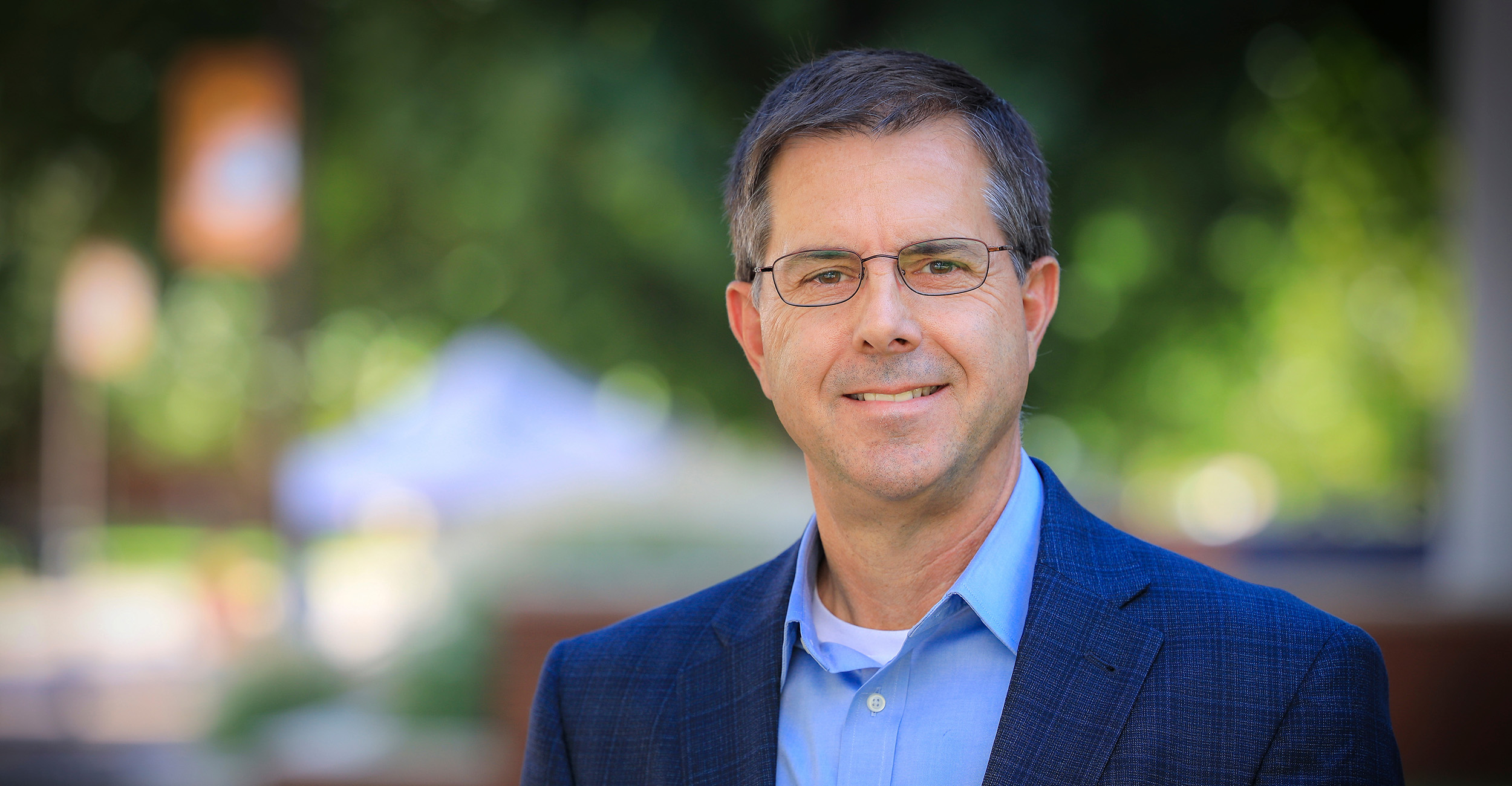 A headshot of Dave Lalman. He is wearing glasses, a blue, collared button-up shirt and a blue blazer. The background is blurred green from trees.