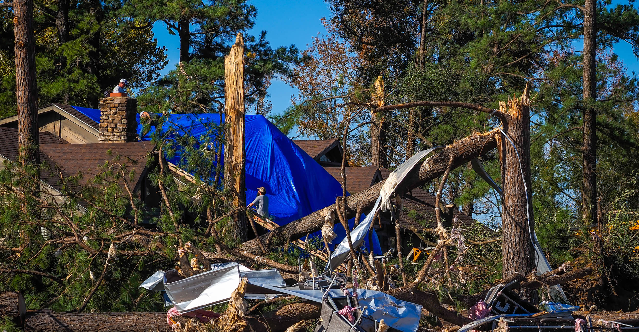 Tornado debris wraps around splintered trees in front of a two-story home partially covered with a large bright blue tarp. Other tall trees tower above the house in the background.