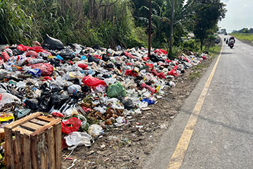 A large pile of illegally dumped trash, including plastic bags, household waste and debris, lines the shoulder of a rural roadside beside tall grass and trees, illustrating the environmental and public safety issues associated with roadside dumping.