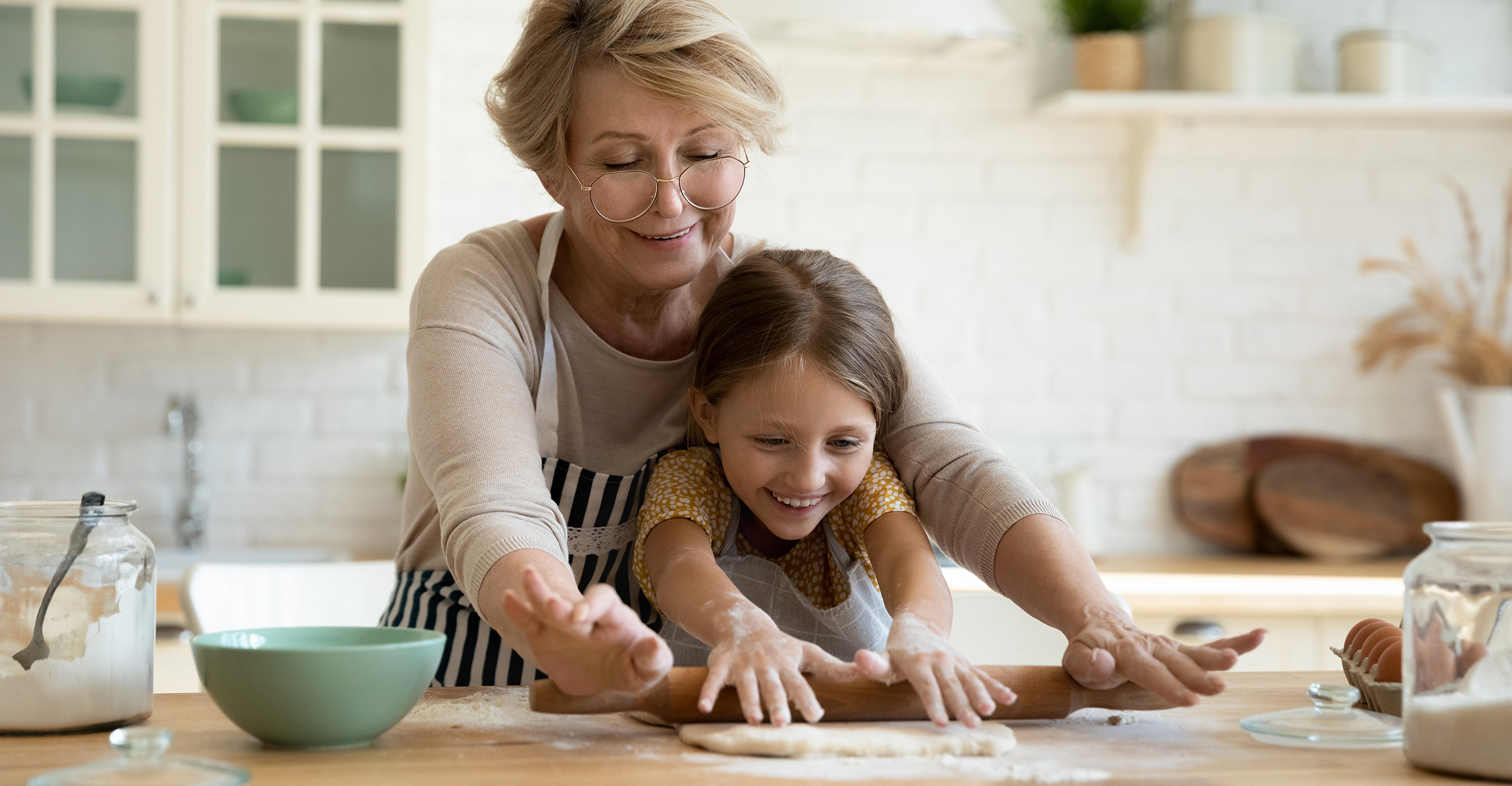 Older woman wearing a striped apron is standing behind a young, brown-haired girl and helping young girl roll out cookie dough with a rolling pin on a wooden counter.