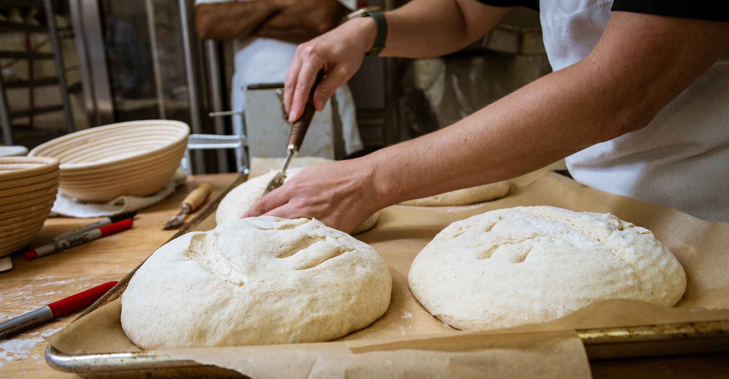 Four loaves of sourdough bread on a baking tray. Human hands use a small kitchen tool to draw lines and designs on the top of the raw dough.