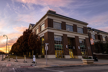 Large, two-story red brick building on the Oklahoma State University campus. There is an orange OSU flag on a black light pole in front of the building and a row of trees along one side of the building.