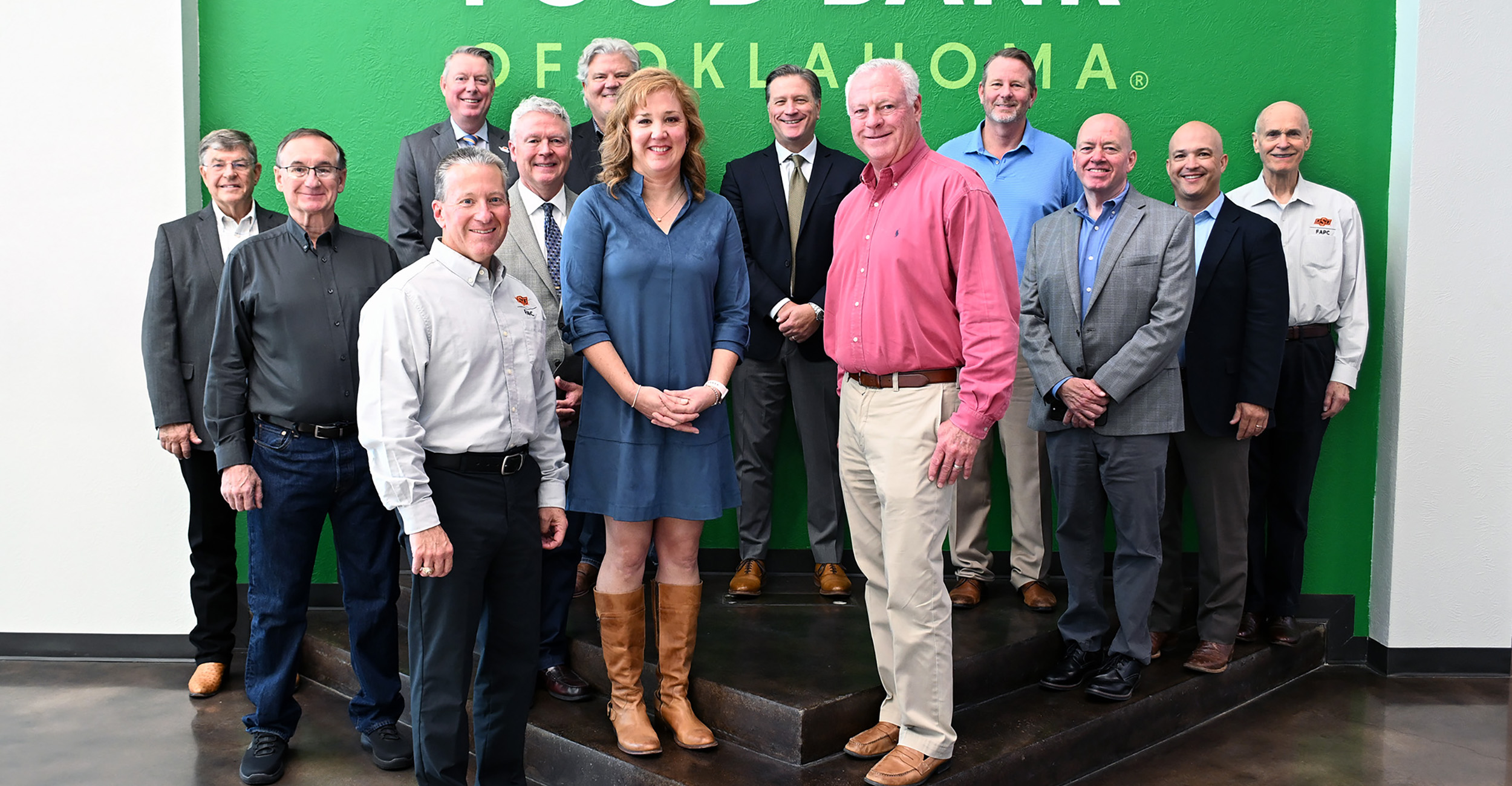 A group of 13 men and women, some standing on a platform, in front of a large, green sign that reads Food Bank of Oklahoma.