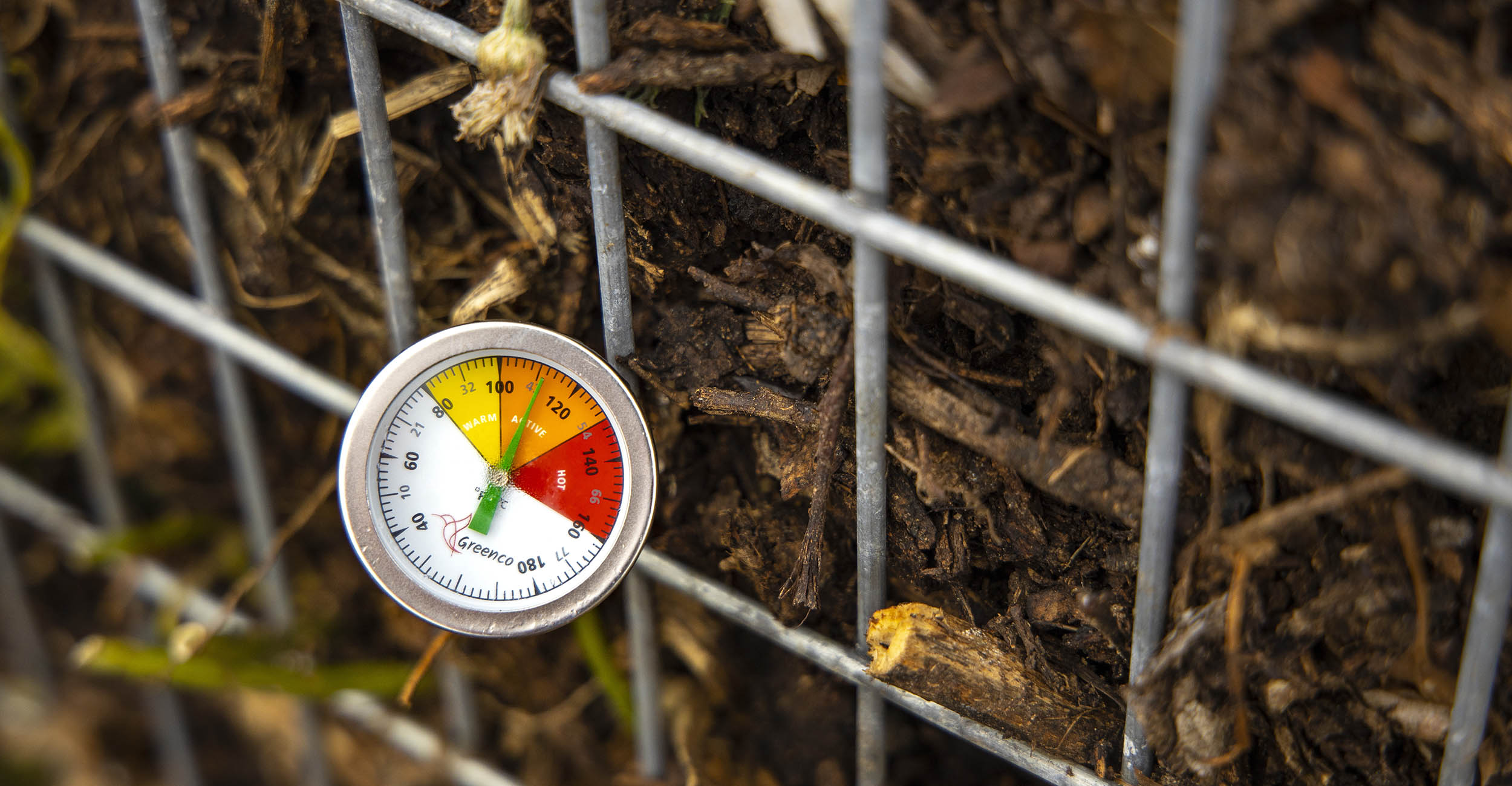 A red, yellow, orange and green temperature gauge stuffed in the side of a metal crate of brown soil and compost.