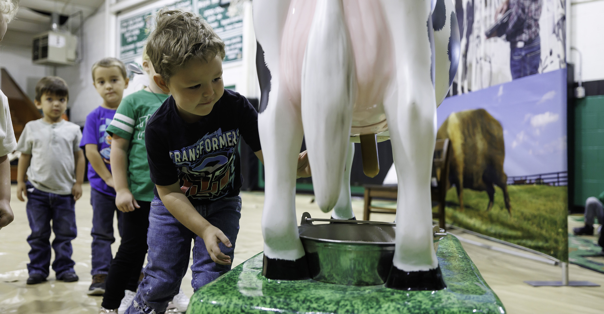 Young boy in black T-shirt milks a life-size plastic cow as other school children wait their turn in line.
