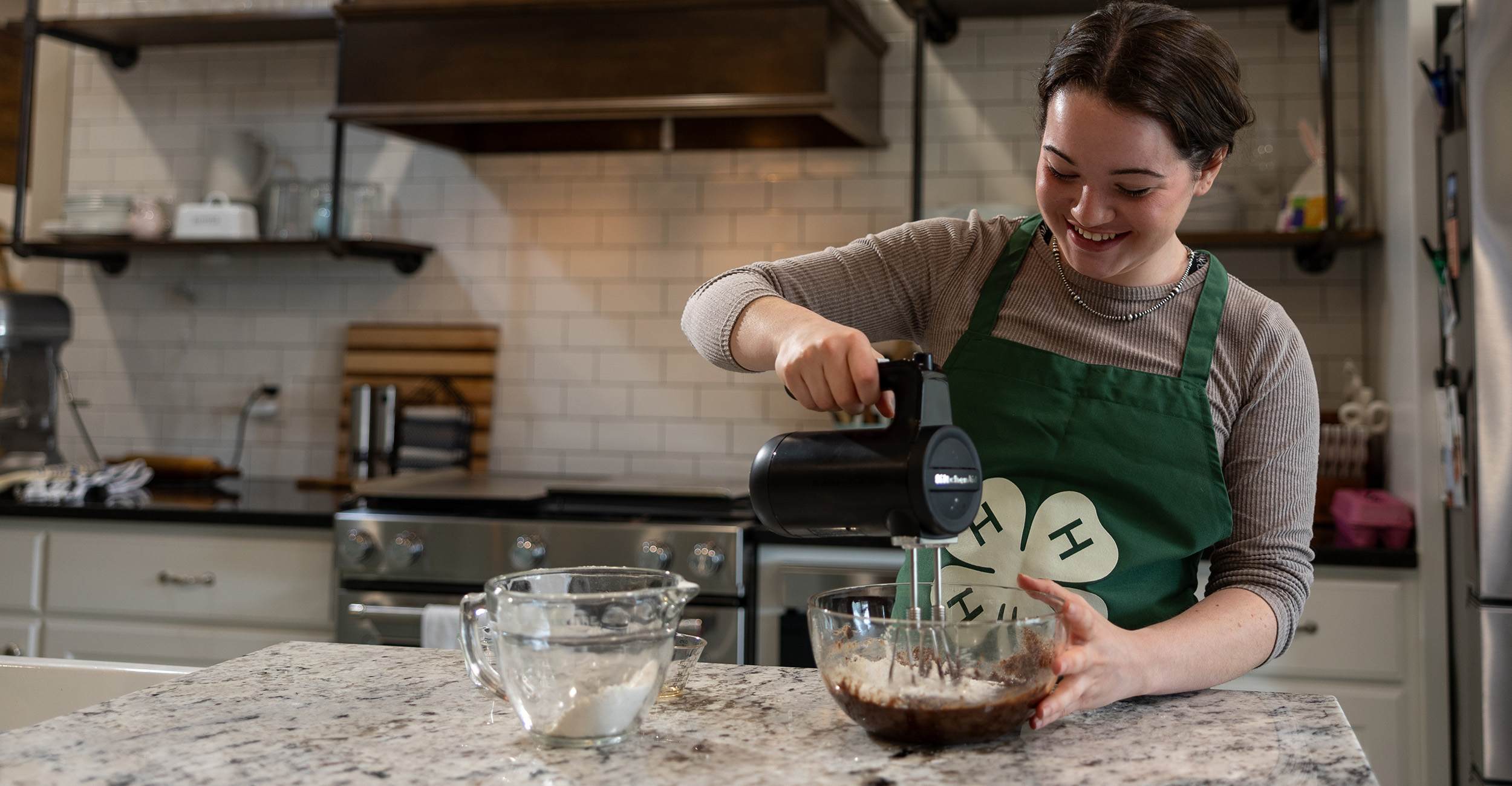 Girl with dark hair in a kitchen using a hand mixer to mix batter in a glass bowl.